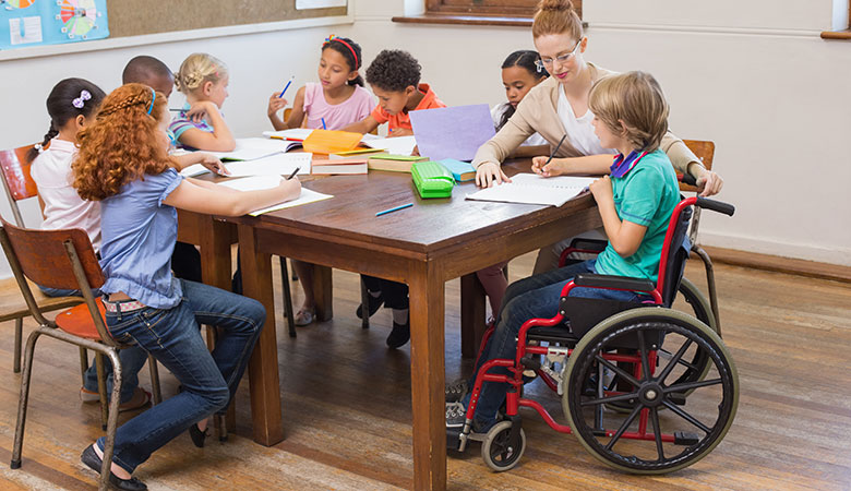 Kinder sitzen über Hefte gebeugt um einen Tisch herum. Dazwischen eine Lehrerin, die sich um einen Schüler mit Rollstuhl kümmert.
