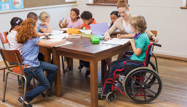 Kinder sitzen über Hefte gebeugt um einen Tisch herum. Dazwischen eine Lehrerin, die sich um einen Schüler mit Rollstuhl kümmert.
