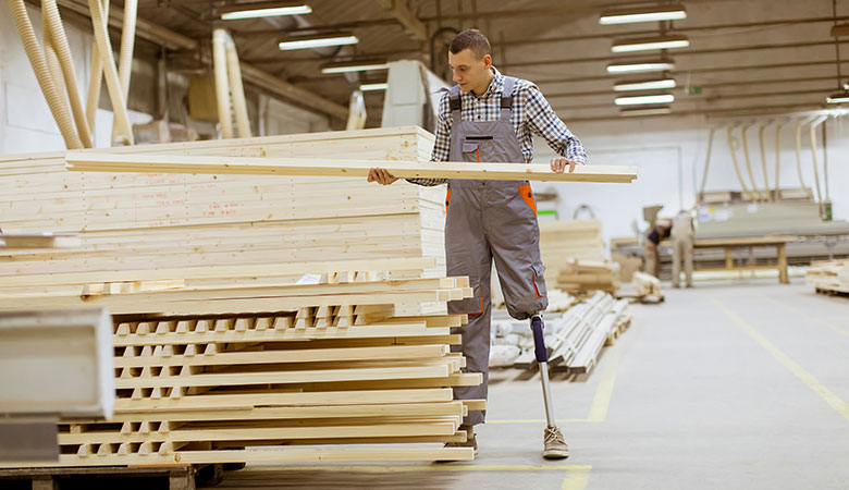 In einer Holzwerkstatt: Mann mit künstlichem Bein hebt Holzbalken von einem Stapel.