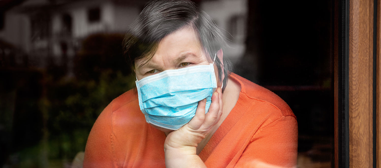 Frau mit Maske sitzt am Fenster, den Kopf auf die Hand gestützt.