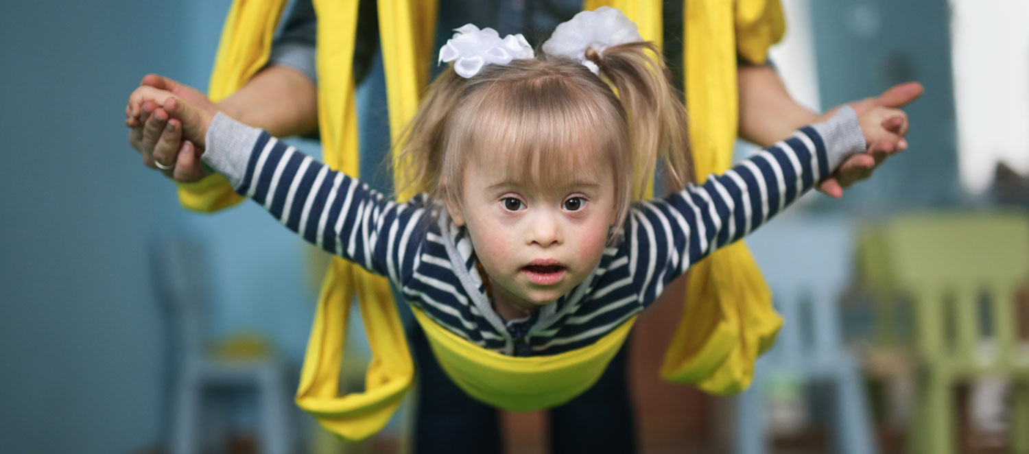 Ein Mädchen hängt in Fliegerstellung in Gymnastik-Bändern und wird von einer Person gehalten.  
