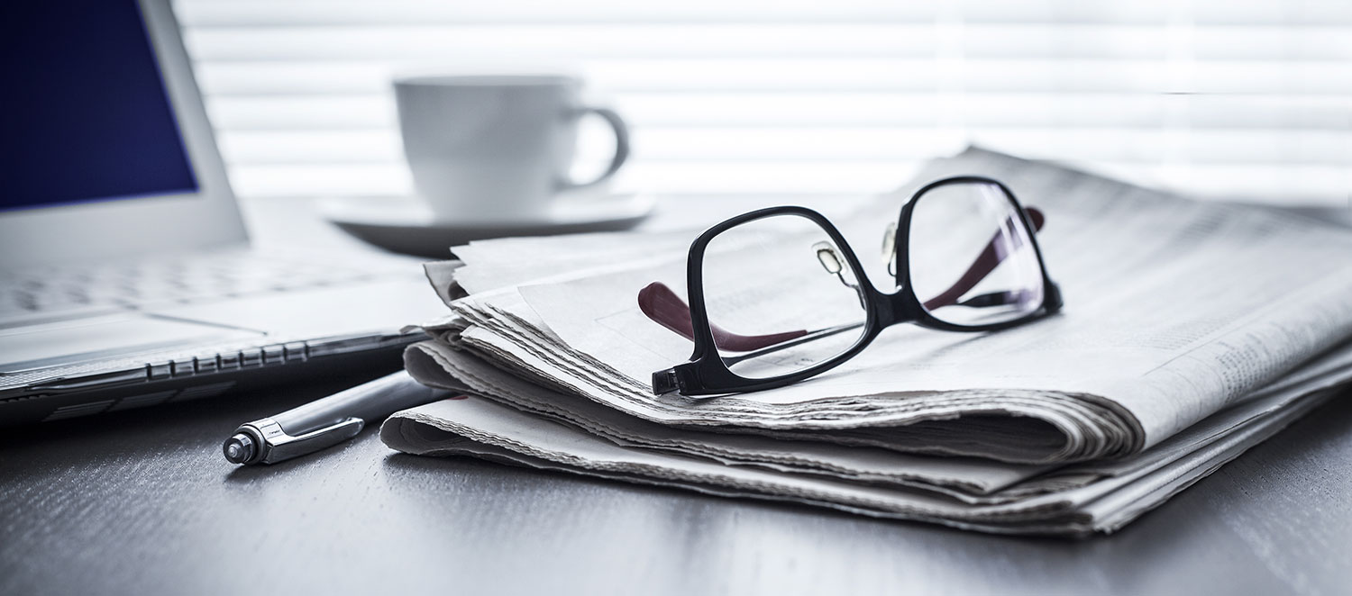 Eine Lesebrille liegt zusammengeklappt auf einer Tageszeitung, daneben ein Stift. Im Hintergrund stehen ein Laptop und eine Kaffeetasse. 